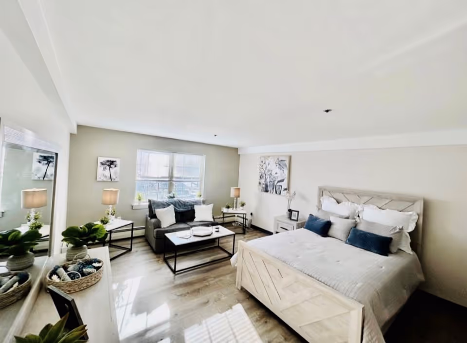 A bright and spacious bedroom featuring a large bed with white and blue pillows, a gray sofa with white cushions, two side tables with lamps, a coffee table, a large mirror, and decorative plants and artwork on the walls.