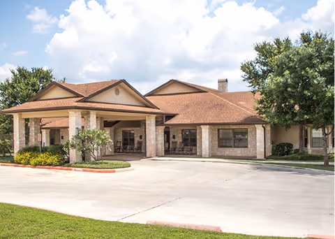 Exterior view of a single-story assisted living facility building with a covered entrance, stone and brick facade, surrounded by greenery and a clear sky with clouds.