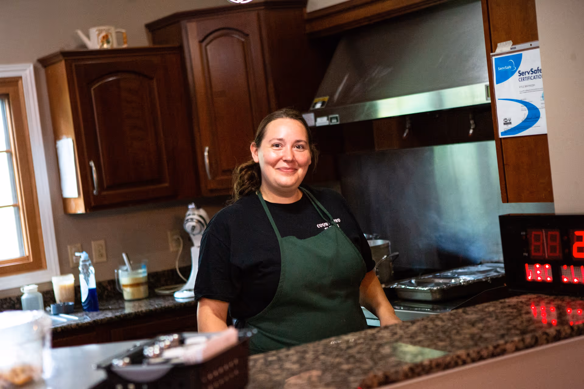 A woman wearing a green apron and black shirt stands behind a granite countertop in a kitchen. Behind her are wooden cabinets, a window, a mixer, and a stainless steel range hood with cooking trays underneath. A ServSafe certification is posted on the cabinet to the right.