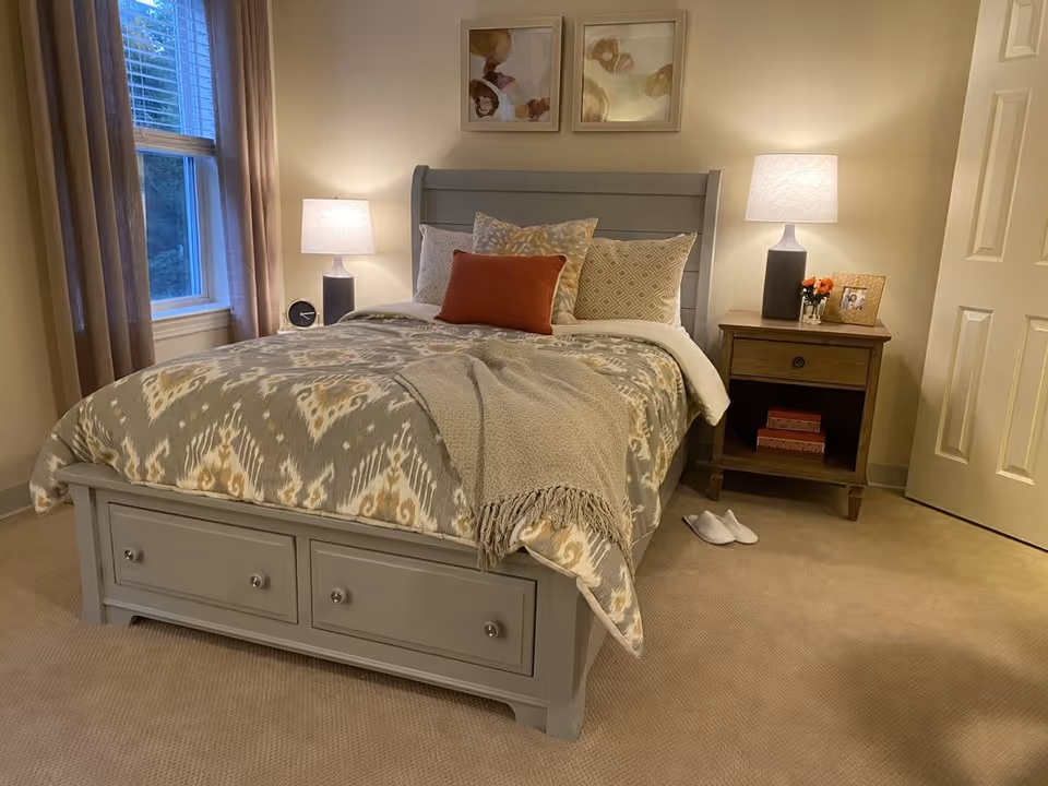 A cozy bedroom with a gray wooden bed frame featuring two drawers at the foot. The bed is made with patterned bedding in neutral tones, accented by a rust-colored pillow and a gray throw blanket. On either side of the bed are nightstands with lamps, one nightstand also holds a small clock and the other has a framed photo and a small flower arrangement. Beige carpet covers the floor, and a window with beige curtains is visible on the left side.