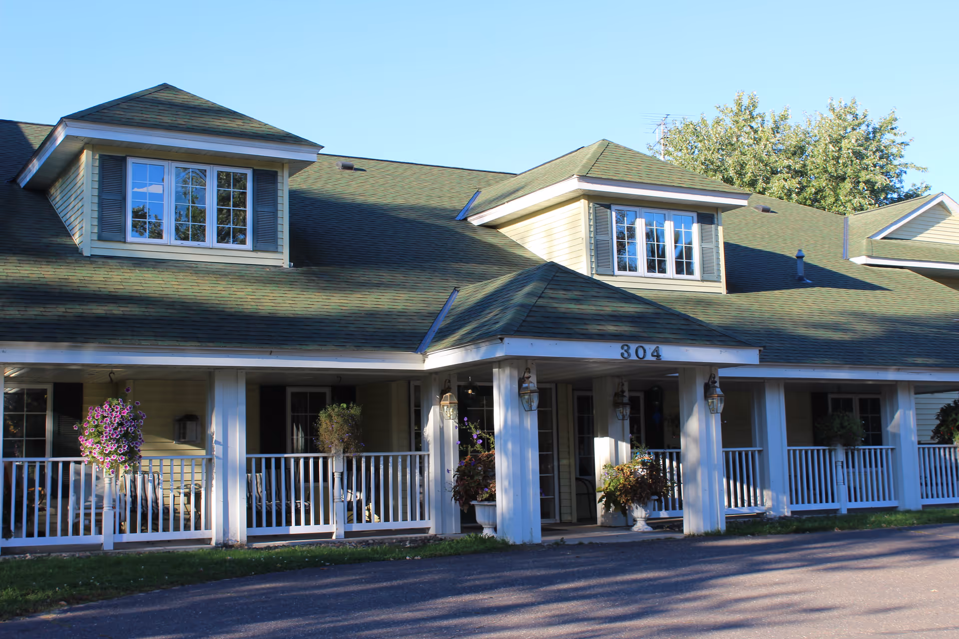 Exterior front view of a senior living facility building with a green roof, beige siding, white railings, and hanging flower baskets. The building number 304 is visible above the entrance.