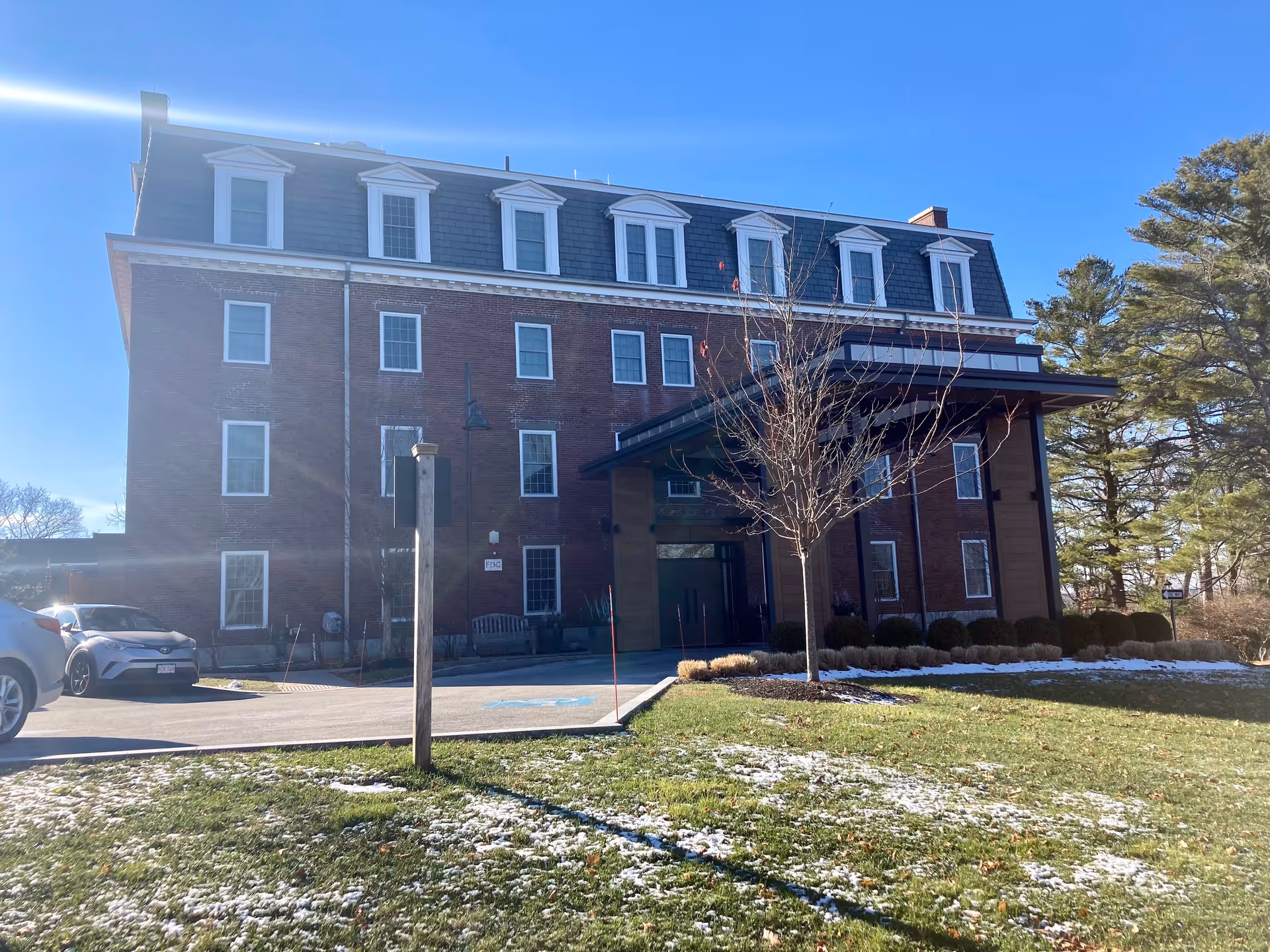 Exterior view of a multi-story brick building with a covered entrance, surrounded by a lawn with patches of snow and some trees under a clear blue sky.