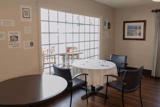 A small dining area with two round tables, one covered with a white tablecloth and surrounded by four black chairs. The room has wooden flooring and beige walls decorated with framed pictures and motivational quotes. Large windows with a grid pattern let in natural light, and an outdoor seating area with wooden chairs and a table is visible through the windows.