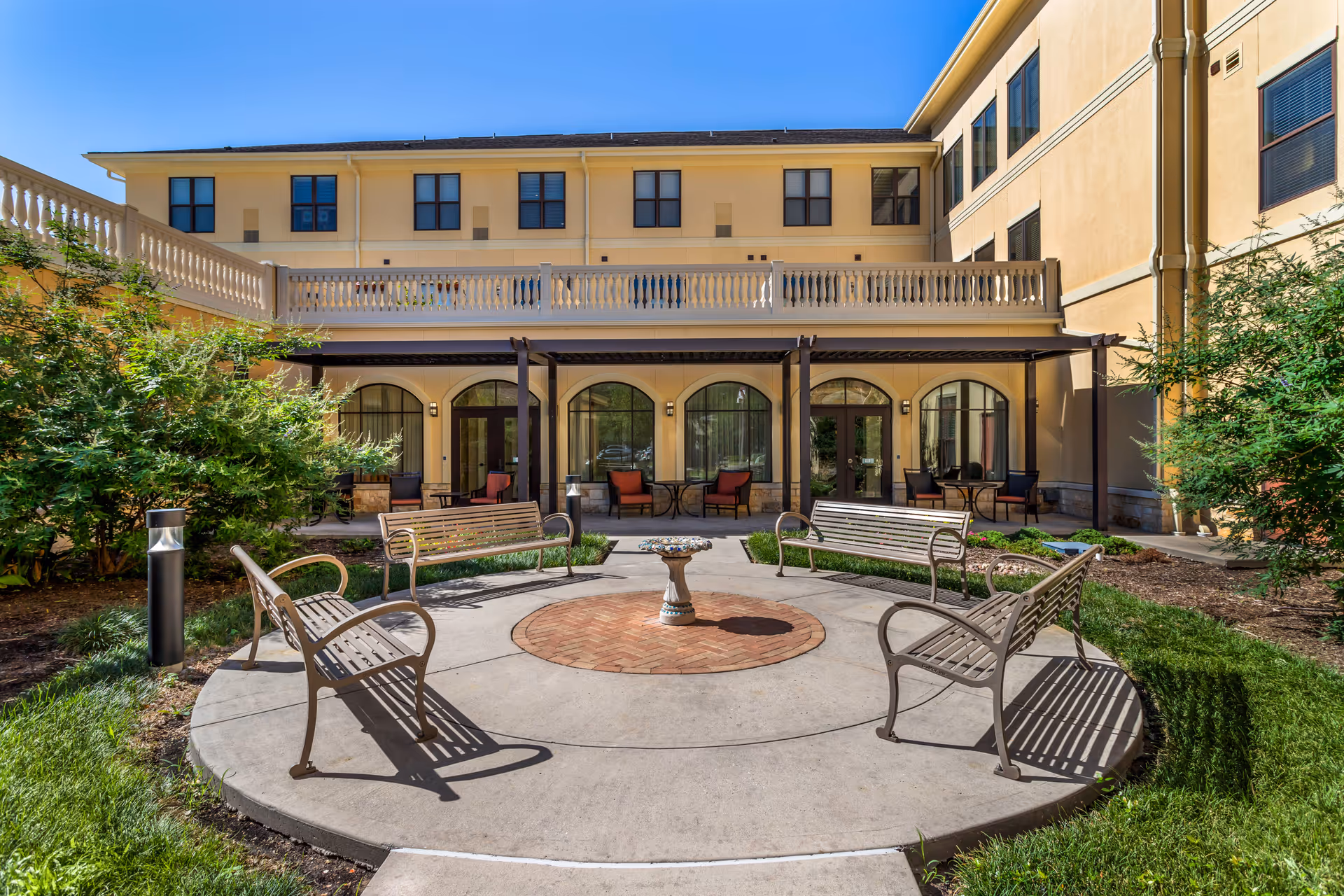 Sunny courtyard with a circular paved seating area, benches, a small fountain, and the surrounding multi-story senior living building.