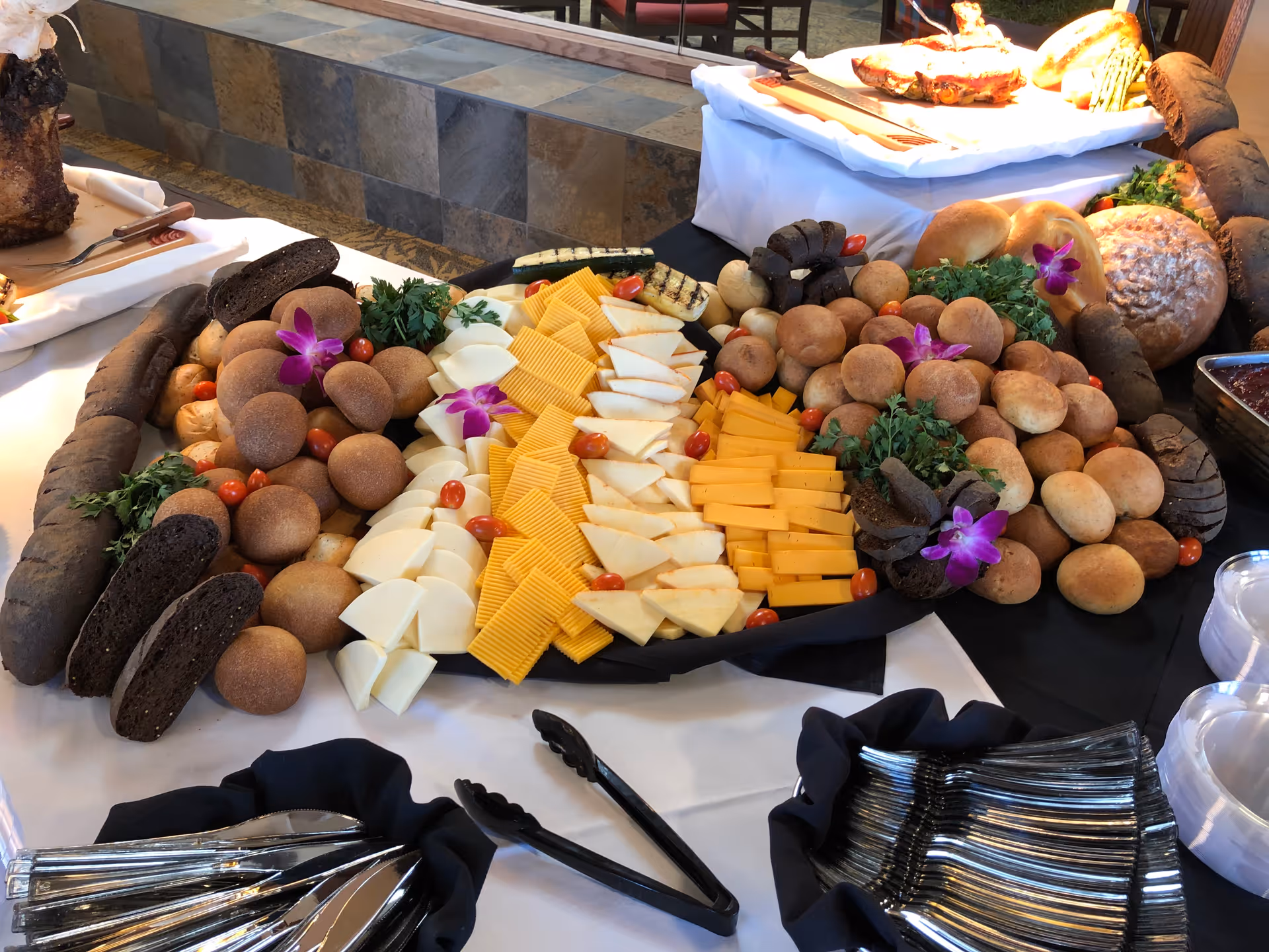 A buffet table with an assortment of sliced cheeses, various types of bread rolls, garnished with cherry tomatoes, parsley, and purple flowers. There are also serving utensils and plates arranged nearby.