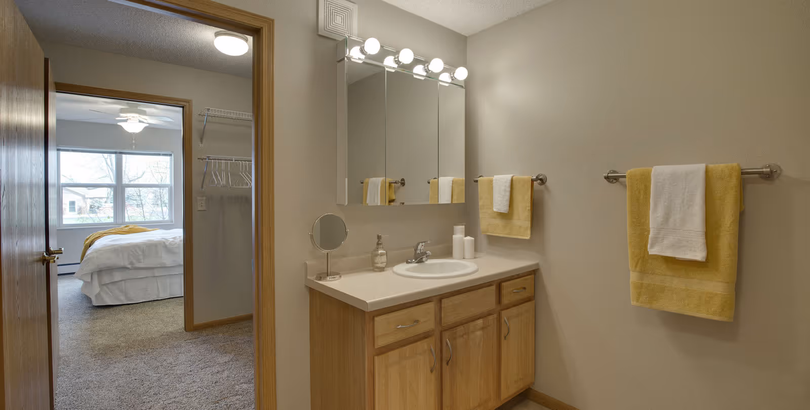 A bathroom vanity area with a sink, wooden cabinets, a large mirror with lights above, and yellow and white towels hanging on a towel rack. The bathroom door is open, showing a carpeted bedroom with a bed, a ceiling fan, and a large window.