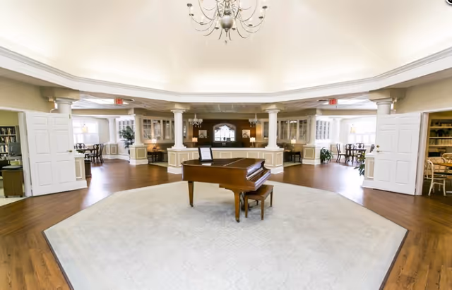 A spacious, elegant common area with a grand piano centered on a large light-colored rug. The room features wooden floors, white columns, and built-in cabinetry with glass doors. There are multiple seating areas with tables and chairs visible through open double doors on either side. A chandelier hangs from the ceiling above the piano.
