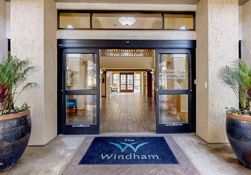 Entrance to The Windham facility showing automatic sliding glass doors, a blue welcome mat with the facility name and logo, two large potted plants on either side of the entrance, and a view into a spacious lobby with wood flooring and seating areas.