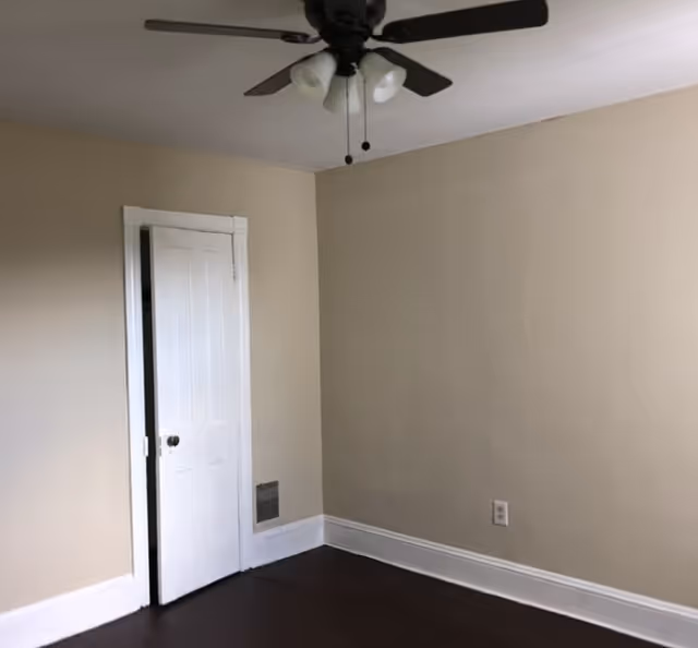 Empty beige room with ceiling fan, a door, and dark hardwood floors.