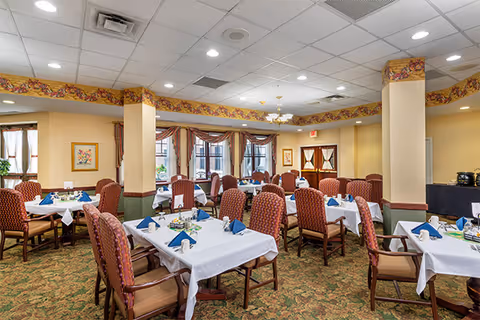 A dining room with multiple tables covered in white tablecloths, each set with blue folded napkins, plates, and condiments. The room has patterned carpet, beige walls with a floral border near the ceiling, and large windows with curtains letting in natural light. Several upholstered chairs with wooden arms surround the tables.