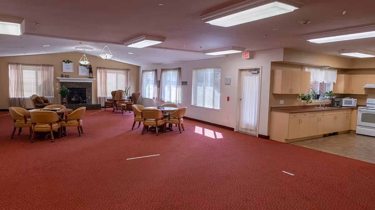 Spacious senior living common room with round dining tables and chairs, a fireplace seating area, and an open kitchenette.