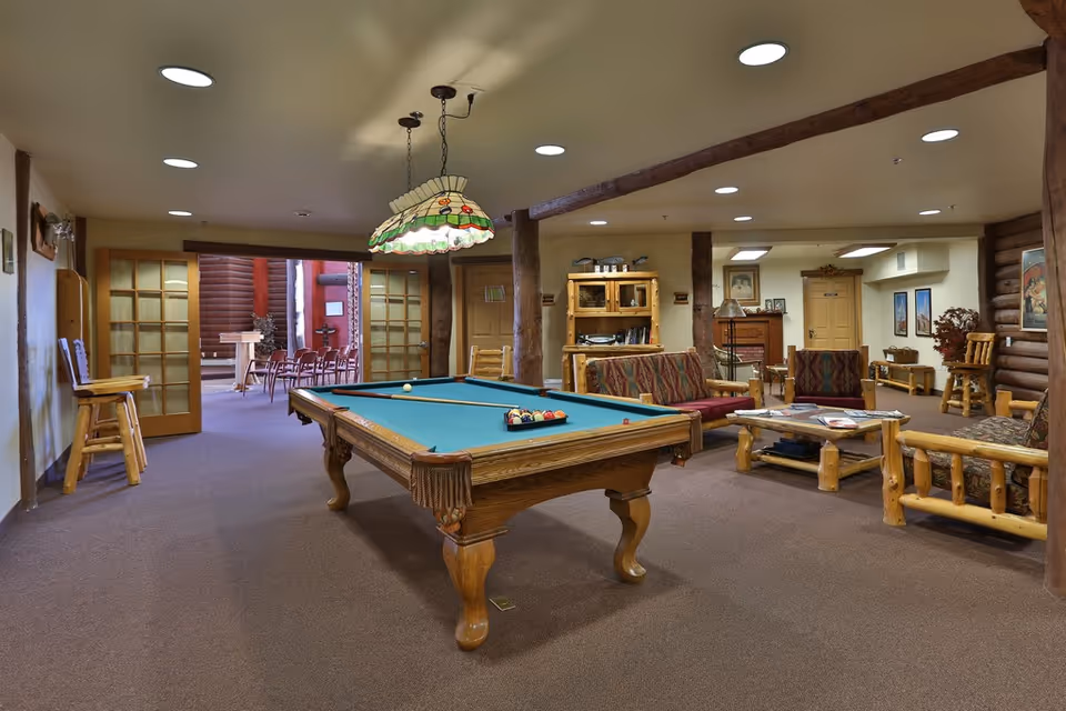 A cozy recreational room with a pool table in the center, surrounded by rustic wooden furniture including sofas and chairs with patterned cushions. The room features wooden beams and log cabin style walls, with a stained glass hanging lamp above the pool table. In the background, there is an open doorway leading to another room with chairs arranged in rows.