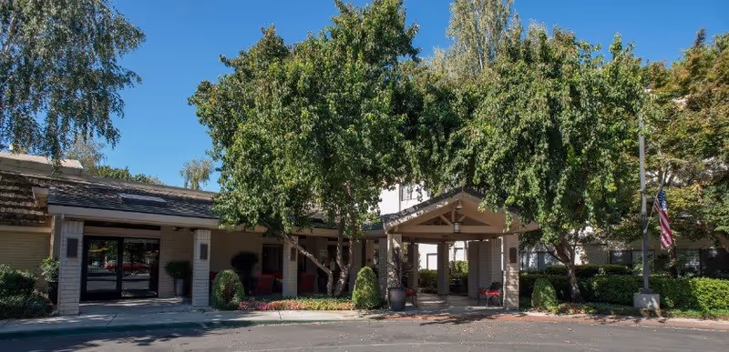 Exterior view of Campus Commons Senior Living facility entrance with a covered drop-off area, surrounded by trees and landscaping under a clear blue sky.