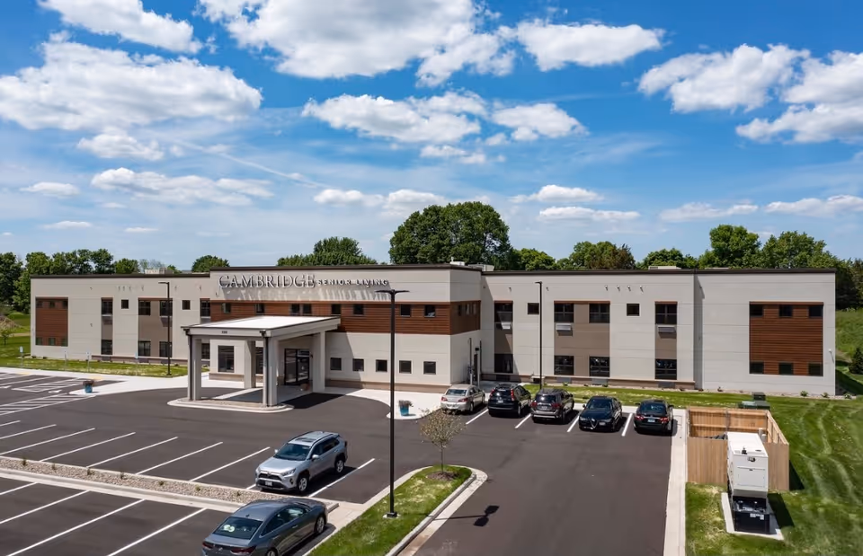 Exterior view of Cambridge Senior Living - Hudson building with a parking lot in front, several cars parked, and a partly cloudy blue sky above.