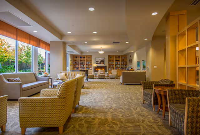 A spacious, well-lit communal lounge with sofas, armchairs, bookshelves and large windows overlooking a garden.