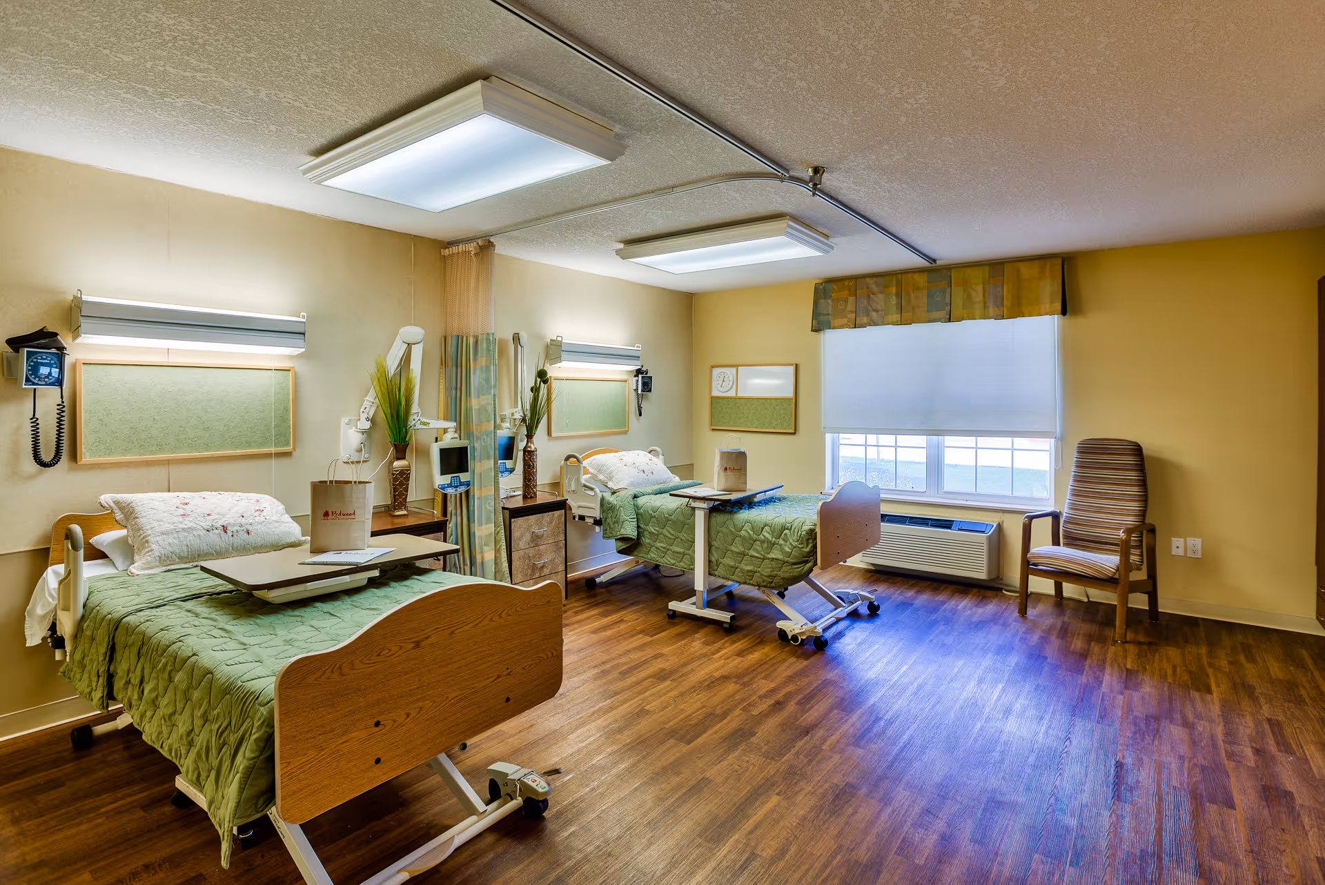 A bright and clean hospital-style room with two adjustable beds, each covered with green quilts and white pillows. Each bed has an overbed table and is positioned against a beige wall with mounted medical equipment and lighting. A privacy curtain is partially visible between the beds. The room has wood flooring, a large window with a white blind and a patterned valance, an air conditioning unit beneath the window, and a striped armchair near the window.