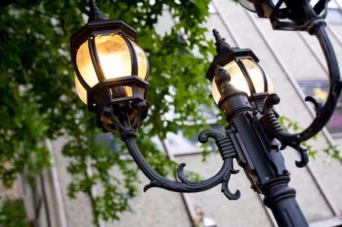 Ornate black outdoor street lamp with glowing lanterns and leafy backdrop.