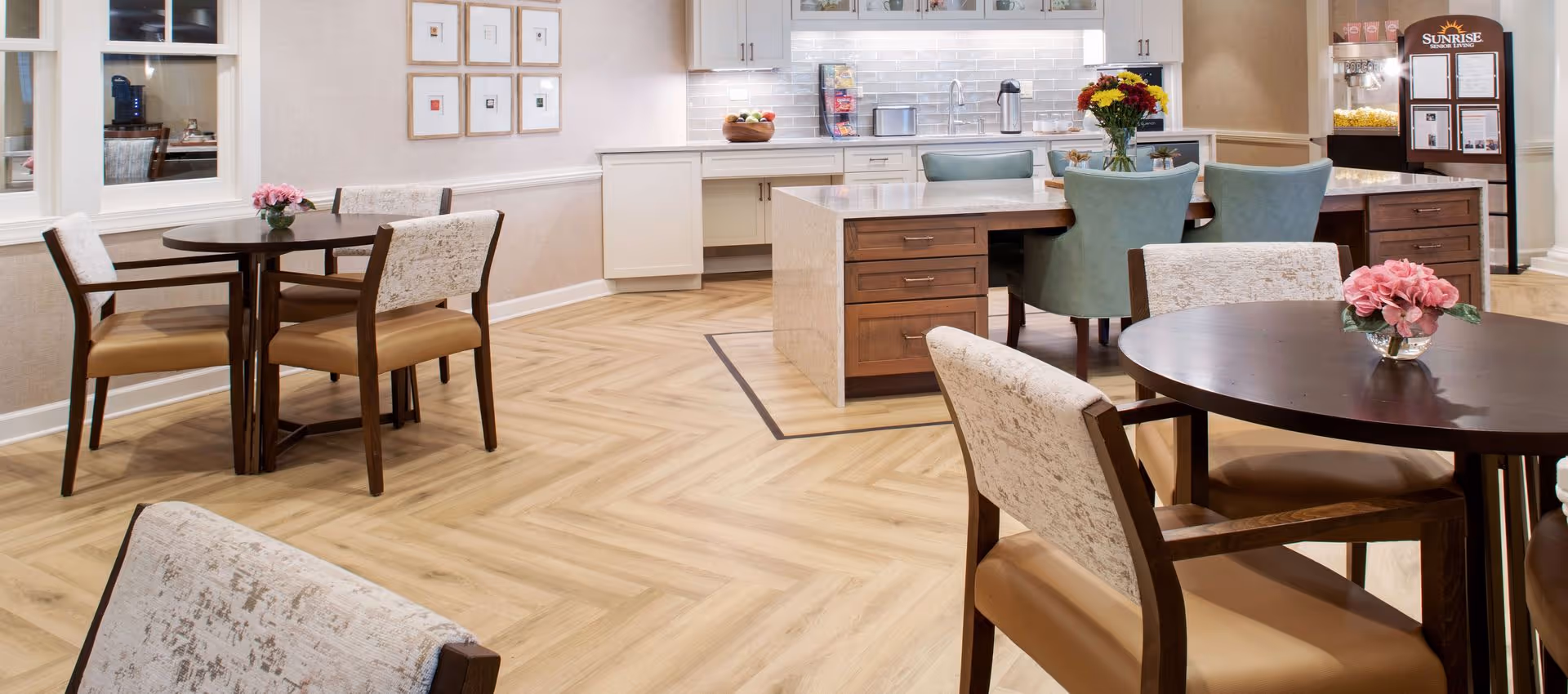 A bright and clean dining area in a senior living facility featuring round wooden tables with cushioned chairs, a kitchen island with teal chairs, a countertop with a fruit bowl and coffee supplies, and a popcorn machine with a Sunrise Senior Living sign in the background.
