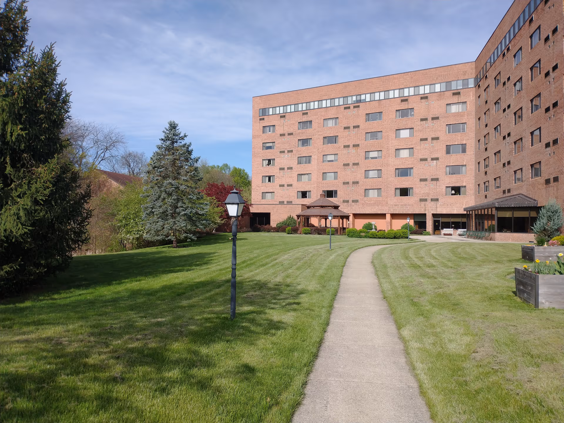A large brick senior living facility building with multiple windows, surrounded by a well-maintained lawn and trees. A concrete pathway leads from the foreground to the building entrance, passing by lampposts and a wooden gazebo. The sky is clear with some light clouds.