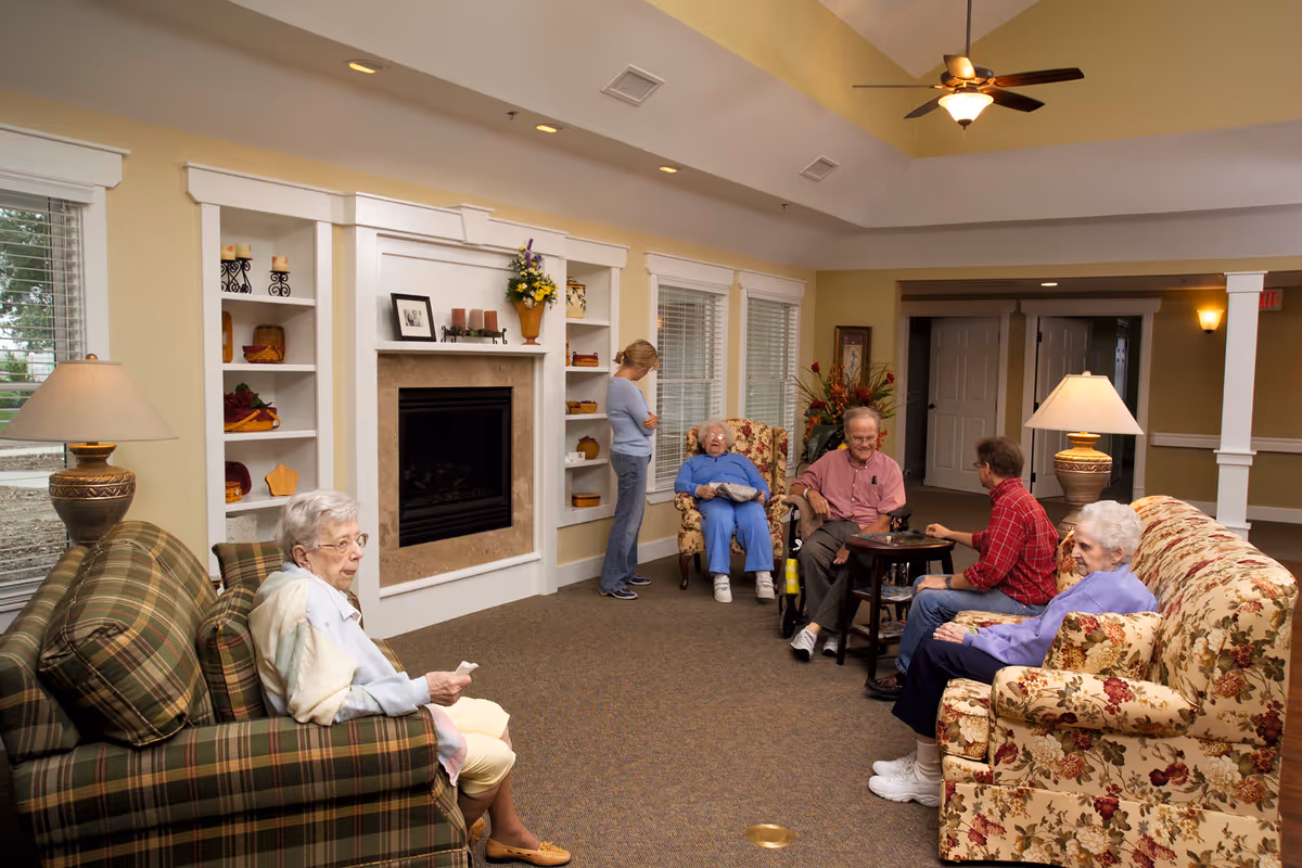 A comfortable senior living lounge with several older adults and a caregiver seated on sofas and chairs near a fireplace and windows.