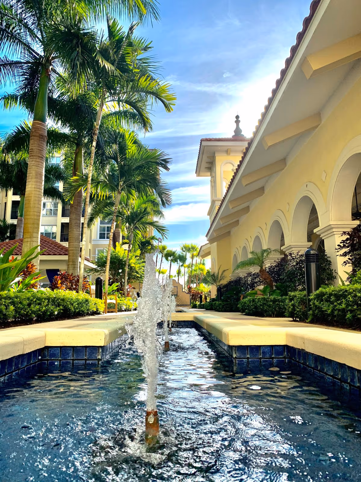 Outdoor view of a senior living facility featuring a water fountain in a narrow pool surrounded by lush greenery and palm trees. The building on the right has a Mediterranean architectural style with arches and a tiled roof under a bright blue sky.