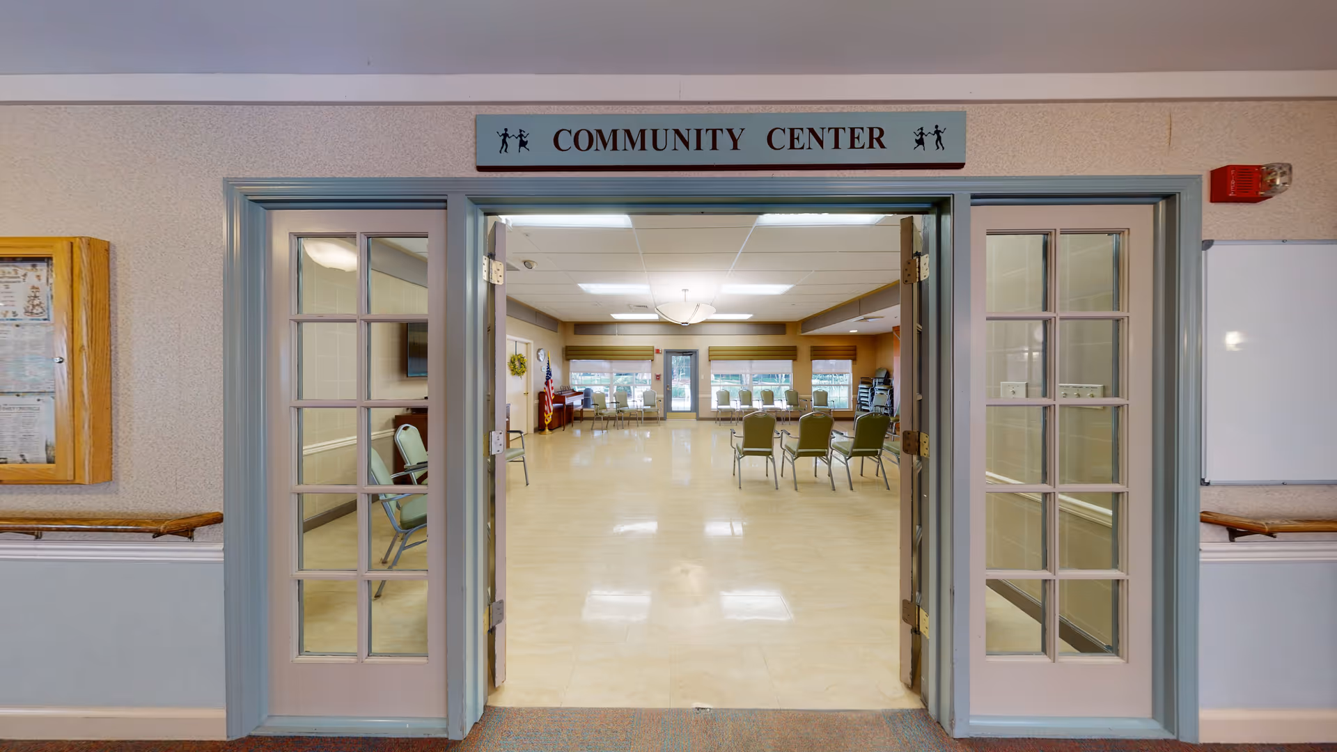 Open double doors with glass panes leading into a community center room with beige tiled floors, several chairs arranged in a semi-circle, large windows with blinds, an American flag, and a piano in the corner.