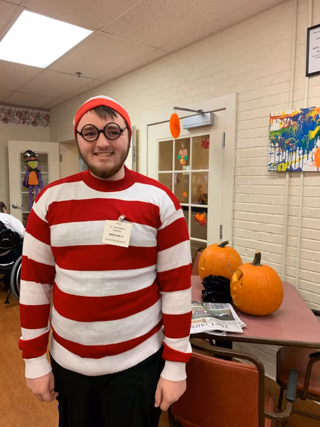 A young man wearing a red and white striped sweater, round black glasses, and a red and white hat, smiling indoors. Behind him is a table with two carved pumpkins and some newspapers. The room has beige brick walls decorated with colorful artwork and autumn-themed decorations on the door and windows.
