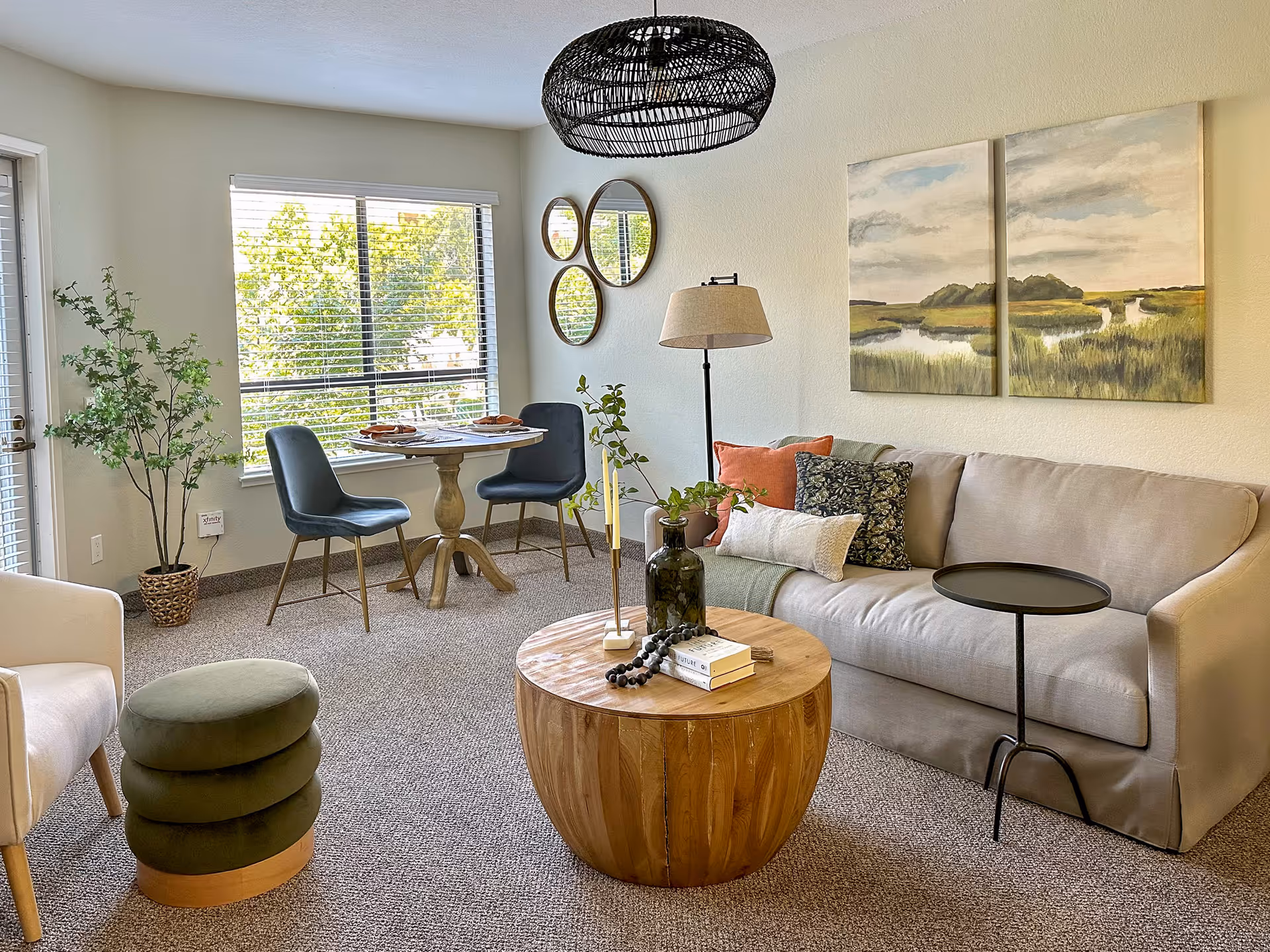 A cozy living room area with a beige sofa adorned with multiple pillows, a round wooden coffee table with decorative items, a floor lamp, and a small black side table. There is a small dining area with a round table and two blue chairs near a window with blinds. The room also features a white armchair, a green ottoman, three round mirrors on the wall, a potted plant, and a large two-panel landscape painting above the sofa.