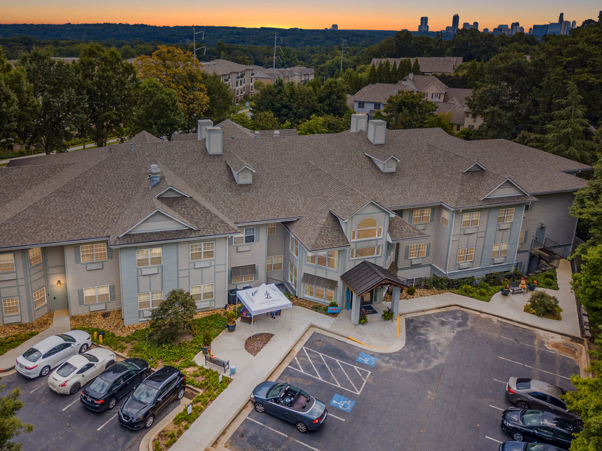 Aerial view of Legacy Ridge at Buckhead senior living facility at sunset, showing a large two-story building with a pitched roof, surrounded by trees and parked cars in the parking lot. The entrance has a covered drop-off area with a canopy displaying the facility's name and logo.