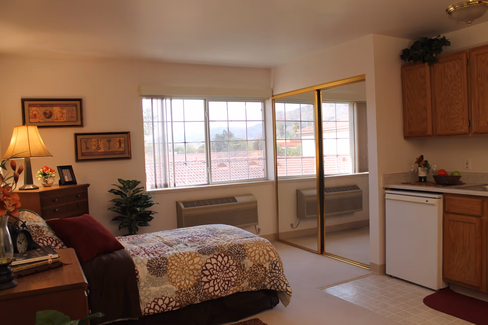 A cozy bedroom with a bed covered in a floral patterned comforter and a burgundy pillow. There is a wooden nightstand with a lamp, a small plant, and framed pictures. A large window with vertical blinds lets in natural light, showing a view of rooftops and distant mountains. To the right, there is a mirrored closet door and a small kitchenette area with wooden cabinets, a countertop, and a mini refrigerator.