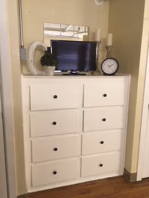 White eight-drawer built-in dresser in an interior alcove topped with a small TV, clock, candles and decorative items.