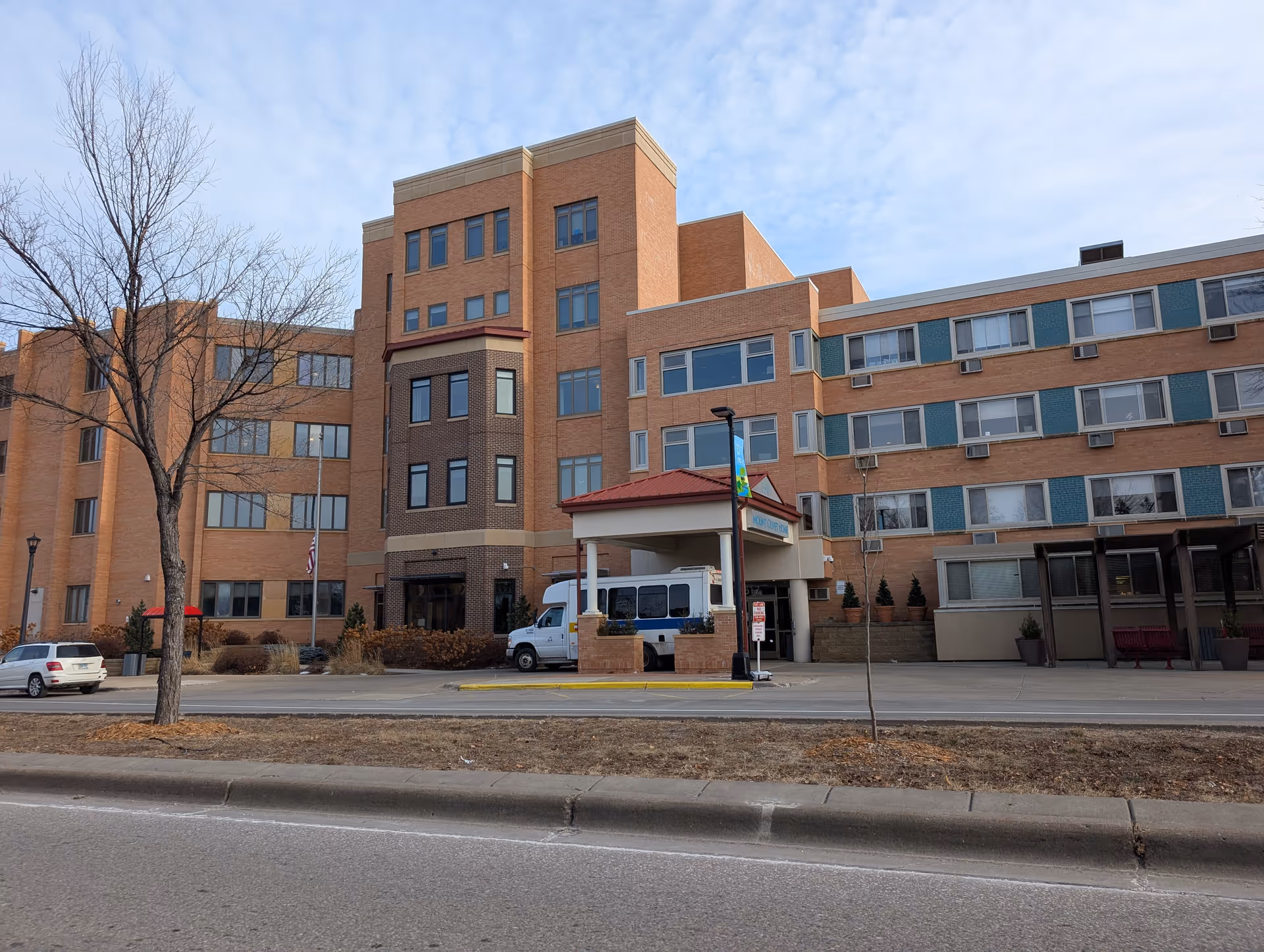 Multi-story brick senior care building with a covered entrance, a parked van, and leafless trees along the street.