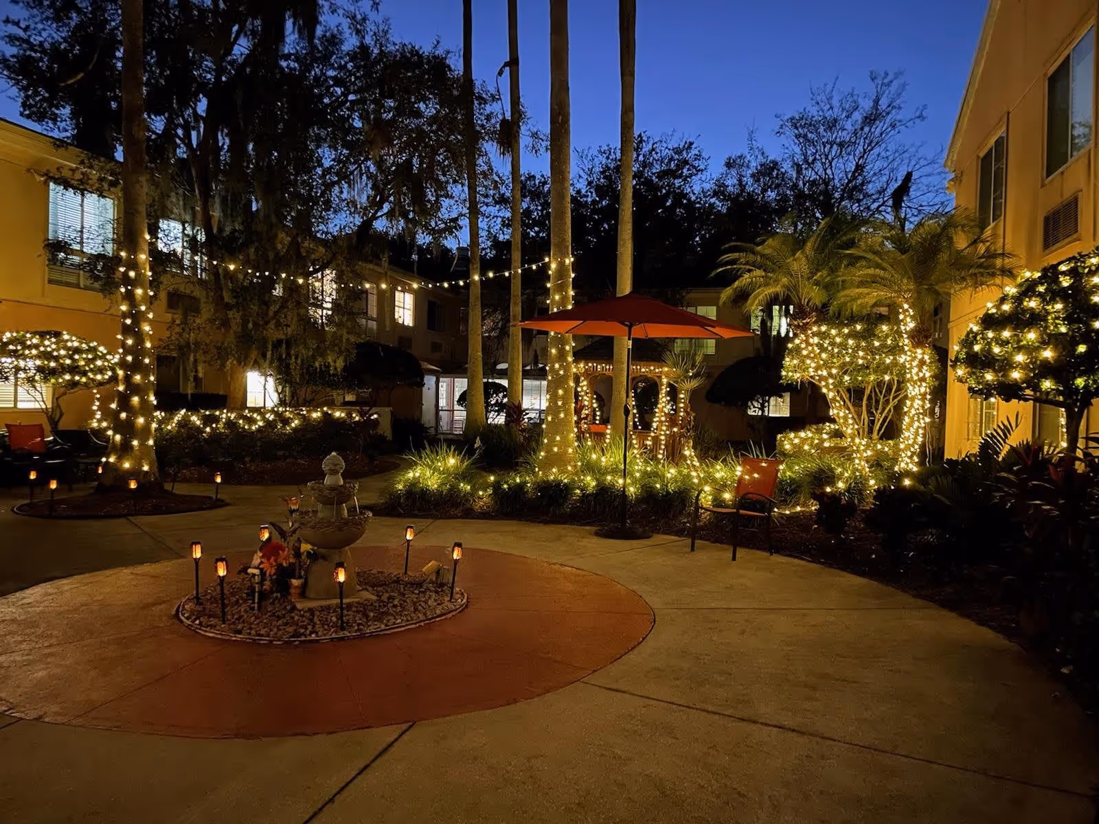 Outdoor courtyard at dusk with trees and bushes wrapped in string lights, a small fountain surrounded by decorative lights, red patio chairs, and a red umbrella. The courtyard is surrounded by a two-story building with lit windows.