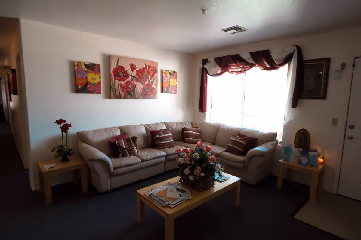 Cozy living room with an L-shaped beige sofa, coffee table with a floral centerpiece and magazines, wall art, and a window with burgundy curtains.