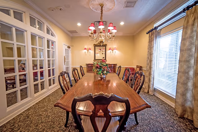 Formal dining room with a long wooden table and chairs, a chandelier overhead, floral centerpiece, mirrored wall and window with curtains.