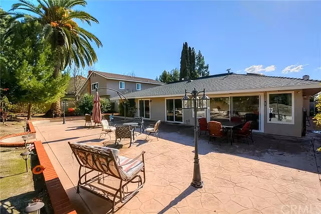 Outdoor patio area of a senior living facility with several chairs and tables arranged on a stamped concrete surface. The patio is adjacent to a single-story building with large windows and sliding glass doors. There are trees and greenery surrounding the patio, including a tall palm tree and other shrubs under a clear blue sky.