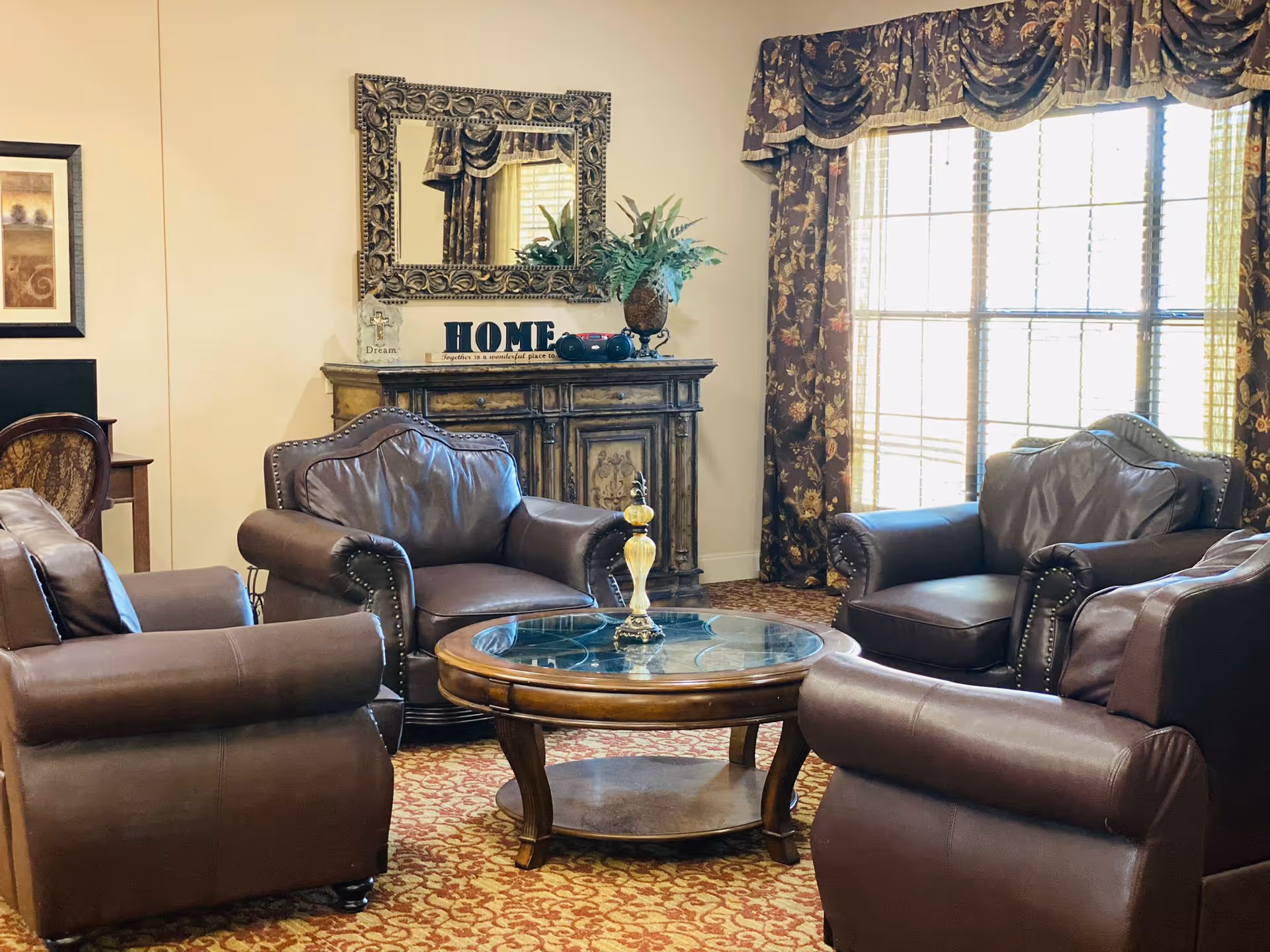 A cozy living room with four brown leather armchairs arranged around a round wooden coffee table with a glass top. Behind the chairs is a wooden cabinet with decorative items including a plant, a mirror with an ornate frame, and a sign that says 'HOME'. The room has a large window with floral curtains letting in natural light.