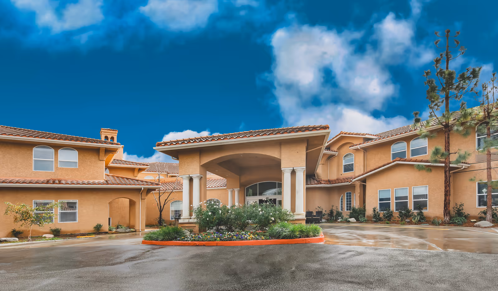 Exterior view of a senior living facility building with a covered entrance supported by columns, surrounded by landscaping and a circular driveway under a partly cloudy blue sky.