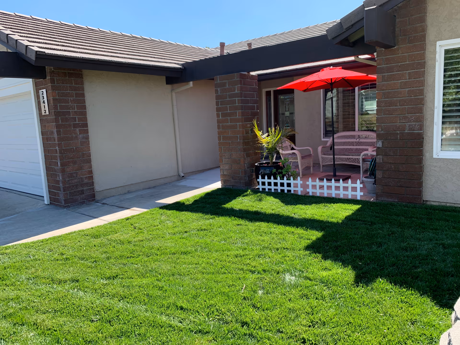 Front porch area of a residential building with a green lawn in the foreground, a small white picket fence, potted plants, a pink bench, two pink chairs, and a red patio umbrella providing shade.