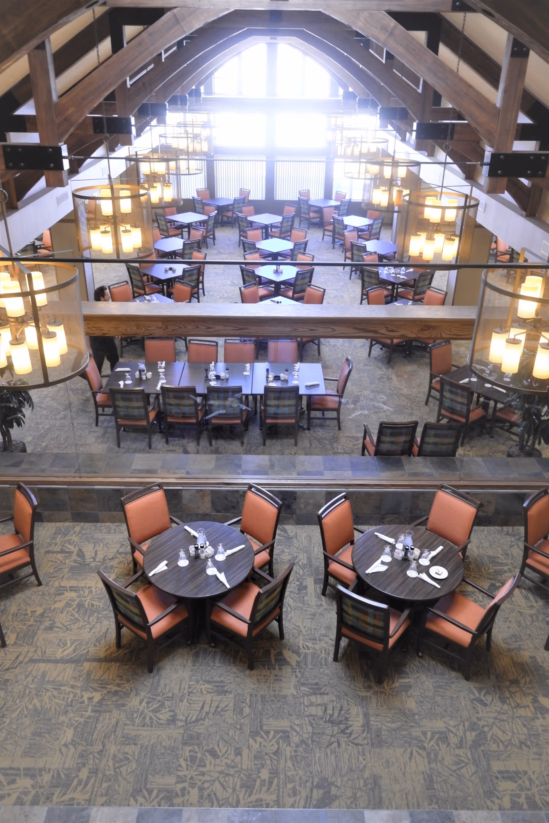 Interior view of a spacious dining room with multiple round and rectangular tables set with napkins and utensils. The room features high vaulted wooden ceilings with large hanging light fixtures and large windows letting in natural light.