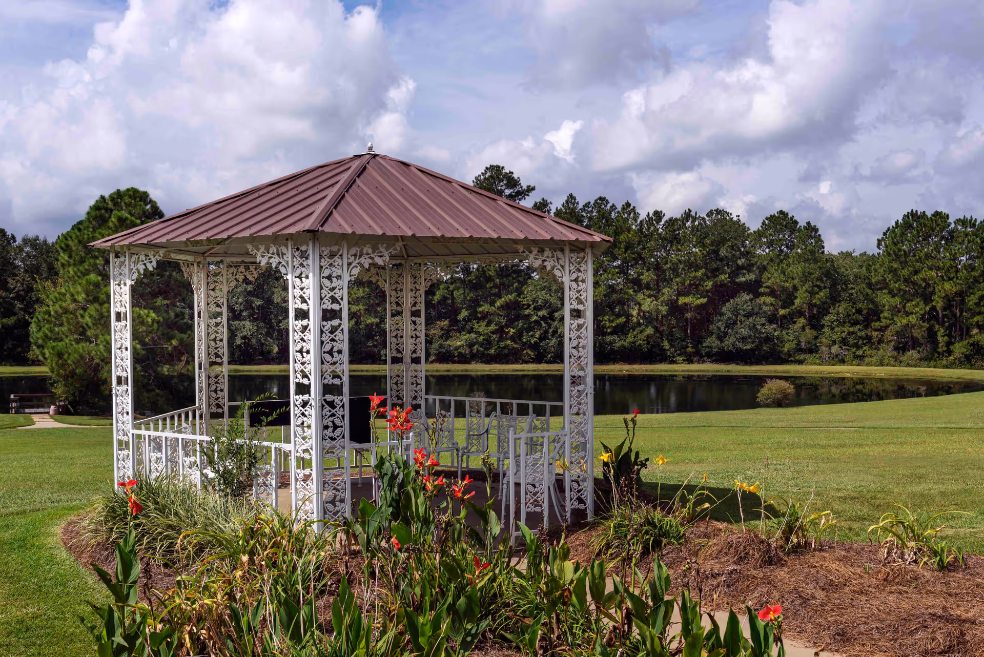 A white metal gazebo with intricate floral designs stands in a garden area with green grass and colorful flowers. Behind the gazebo is a calm pond surrounded by trees under a partly cloudy sky.