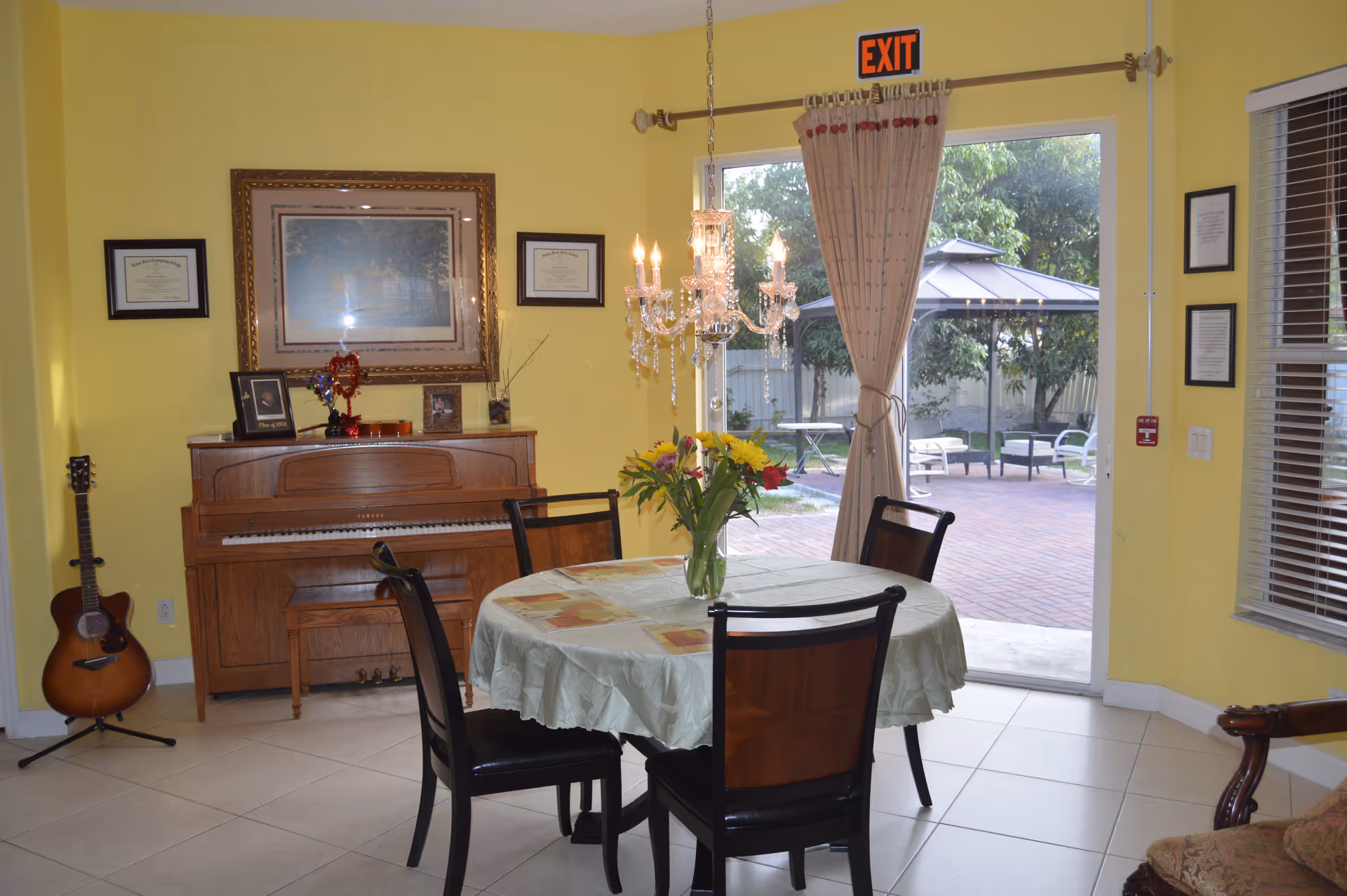A bright room with yellow walls featuring a round dining table covered with a light green tablecloth and four chairs. A vase with colorful flowers is on the table. Behind the table is a wooden piano with framed pictures and certificates on the wall above it. A guitar stands on a stand in the corner. A chandelier hangs above the table. A sliding glass door with beige curtains leads to an outdoor patio area with a gazebo and seating. An exit sign is above the door.