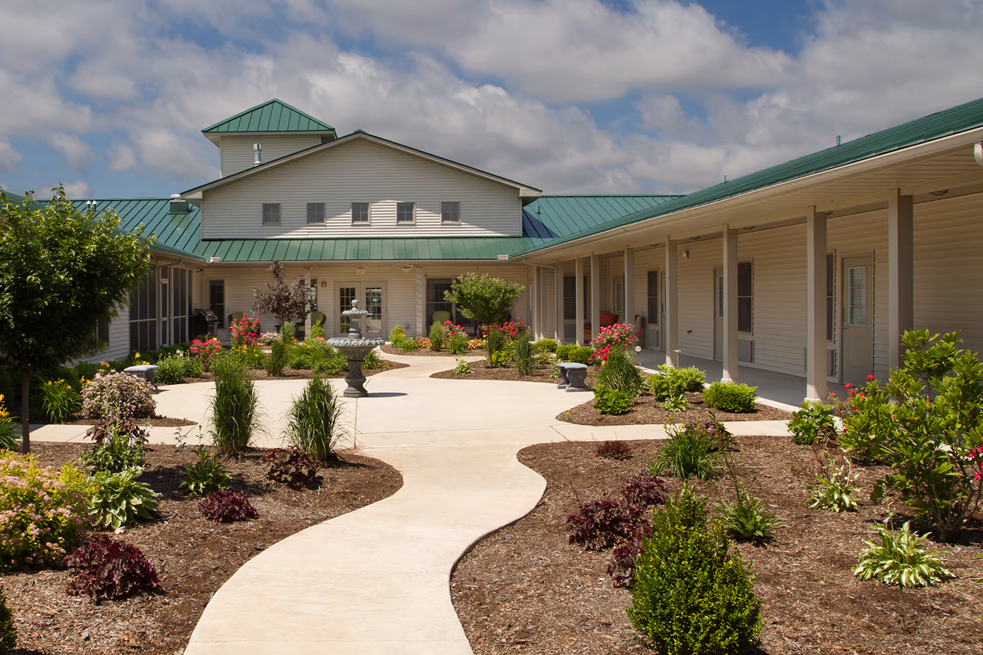 Outdoor courtyard area of Carriage Crossing Senior Living of Arcola featuring a winding concrete pathway surrounded by landscaped garden beds with various shrubs and flowers, leading to a building with a green metal roof under a partly cloudy sky.