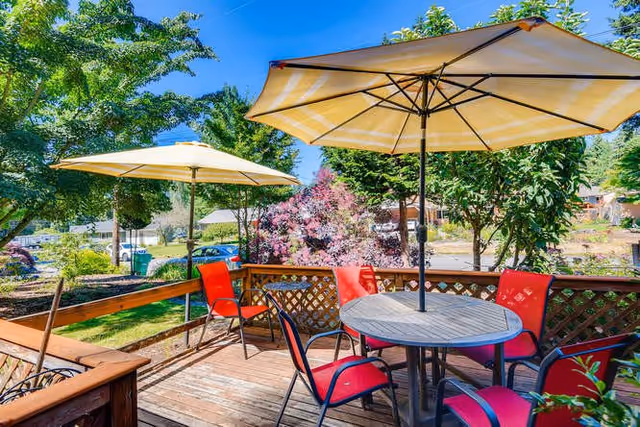 Outdoor wooden deck with a round table and four red chairs under a large beige patio umbrella. Another beige umbrella is visible in the background. The deck overlooks a garden with trees, shrubs, and a clear blue sky.