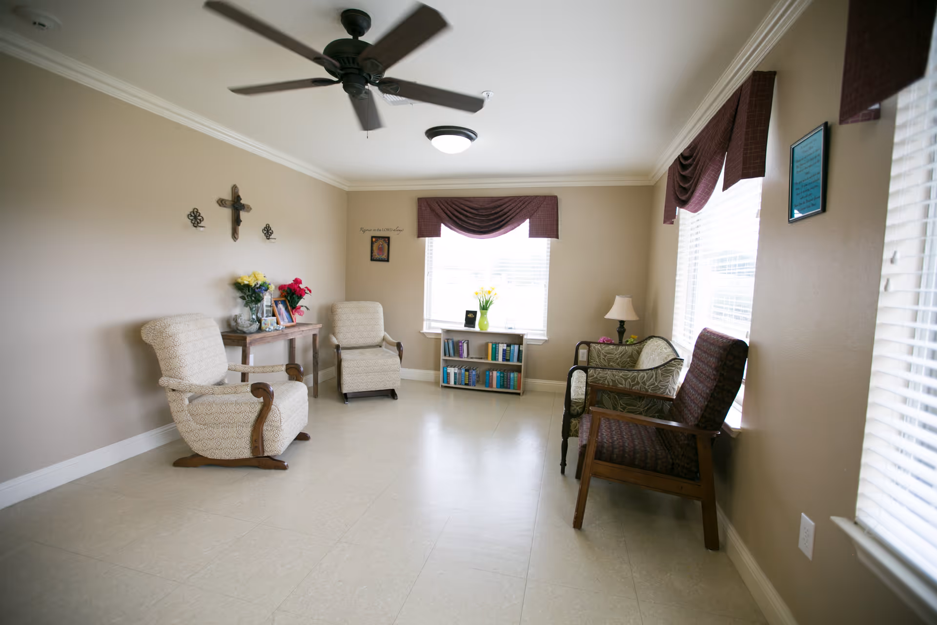 A bright and cozy sitting room with beige walls and tiled floor. The room features two beige upholstered armchairs with wooden armrests on the left, a small wooden table with flowers and framed photos, a bookshelf filled with books under a window with a purple valance, and two additional chairs with patterned upholstery on the right. A ceiling fan and light fixture are mounted on the white ceiling. The room is decorated with a cross and small wall hangings.