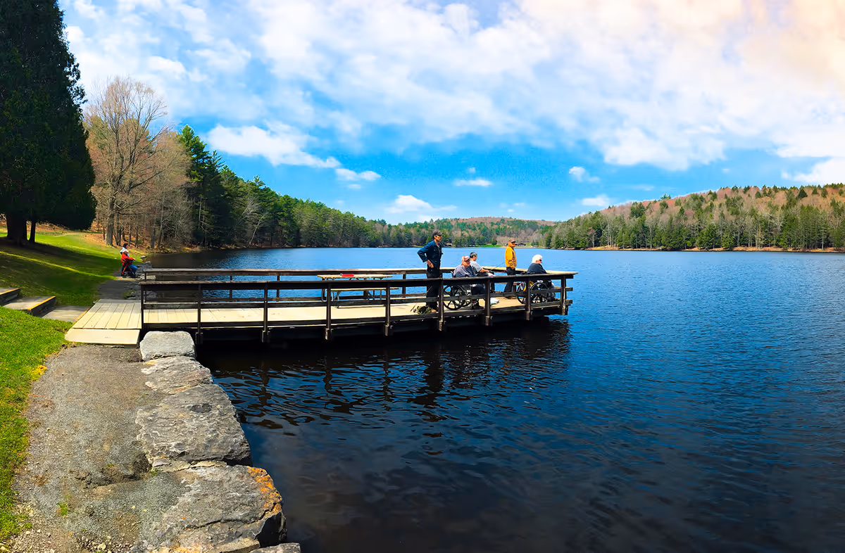 A wooden dock extending over a calm lake surrounded by trees under a partly cloudy sky. Several people, including some in wheelchairs, are on the dock enjoying the view. A person is sitting on a bench near the shore on the left side of the image.