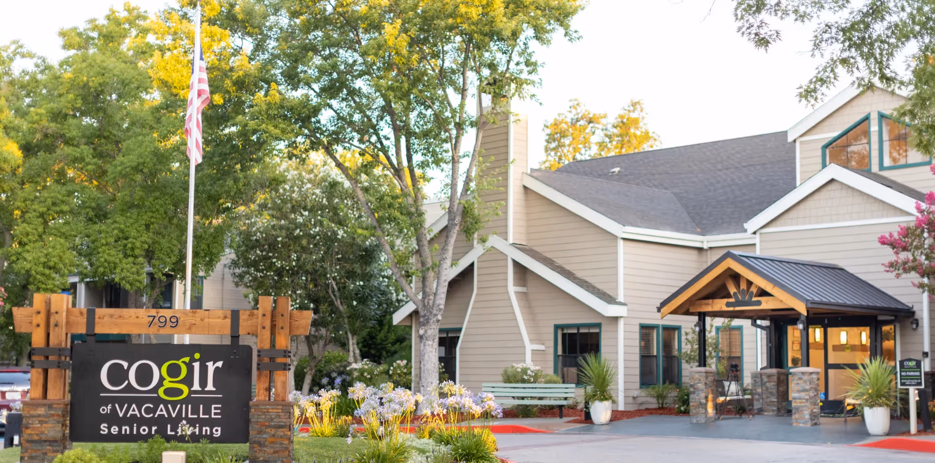 Exterior view of Cogir of Vacaville Senior Living facility with a wooden sign displaying the name and address number 799. The building has beige siding, a dark roof, and a covered entrance supported by stone pillars. There are trees, flowering plants, and a bench in the landscaped area around the entrance.