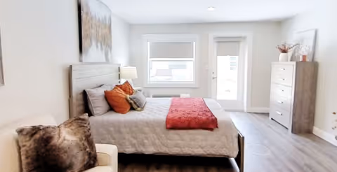 A bright bedroom with a large bed featuring white bedding, orange and gray pillows, and a red throw blanket. There is a white armchair with a furry pillow in the foreground, a wooden dresser with decorative items on top, a window, and a glass door leading outside. The room has light-colored walls and wood flooring.
