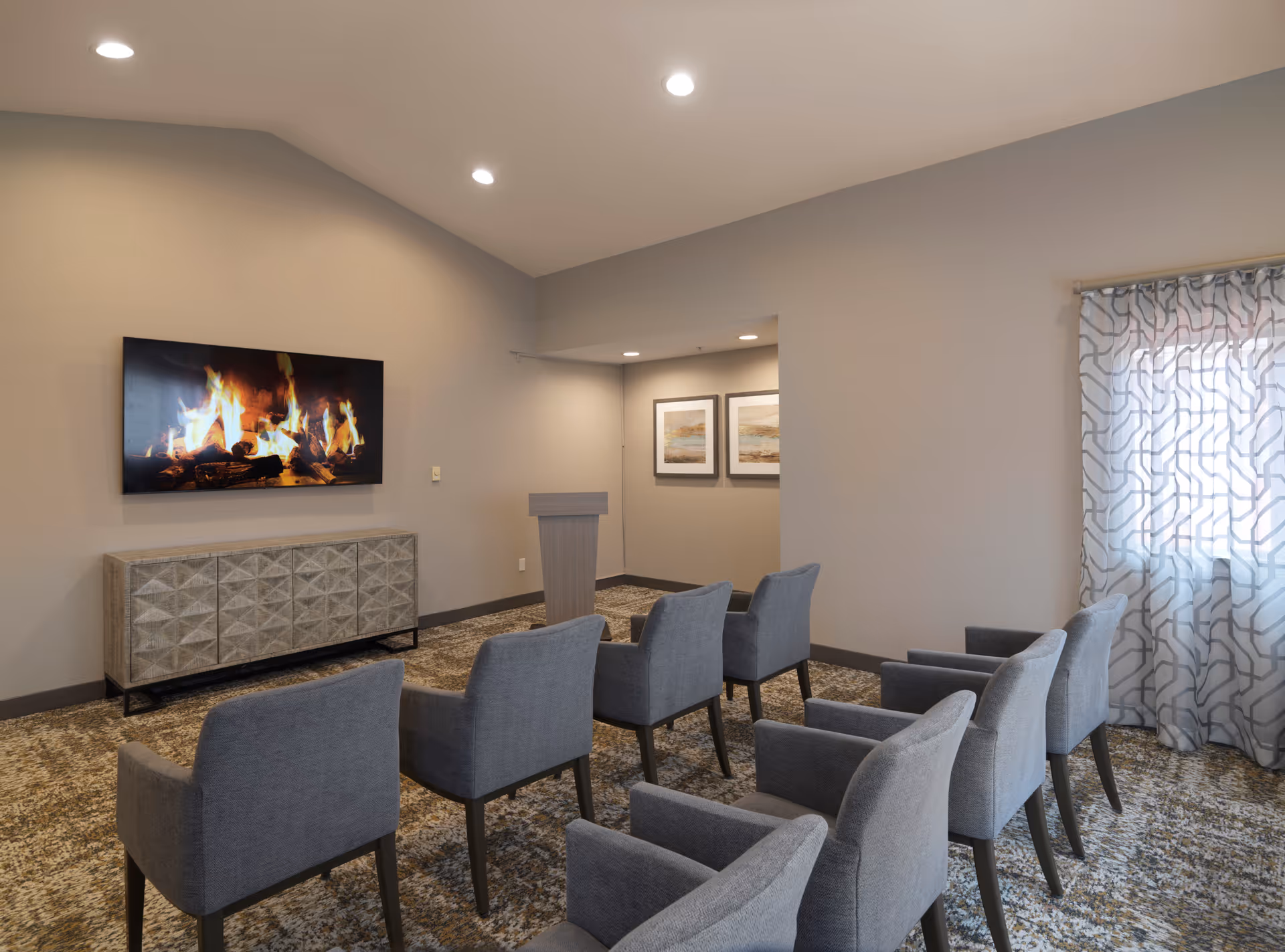 Small meeting room with rows of gray upholstered chairs facing a wall-mounted TV showing a fireplace and a podium.
