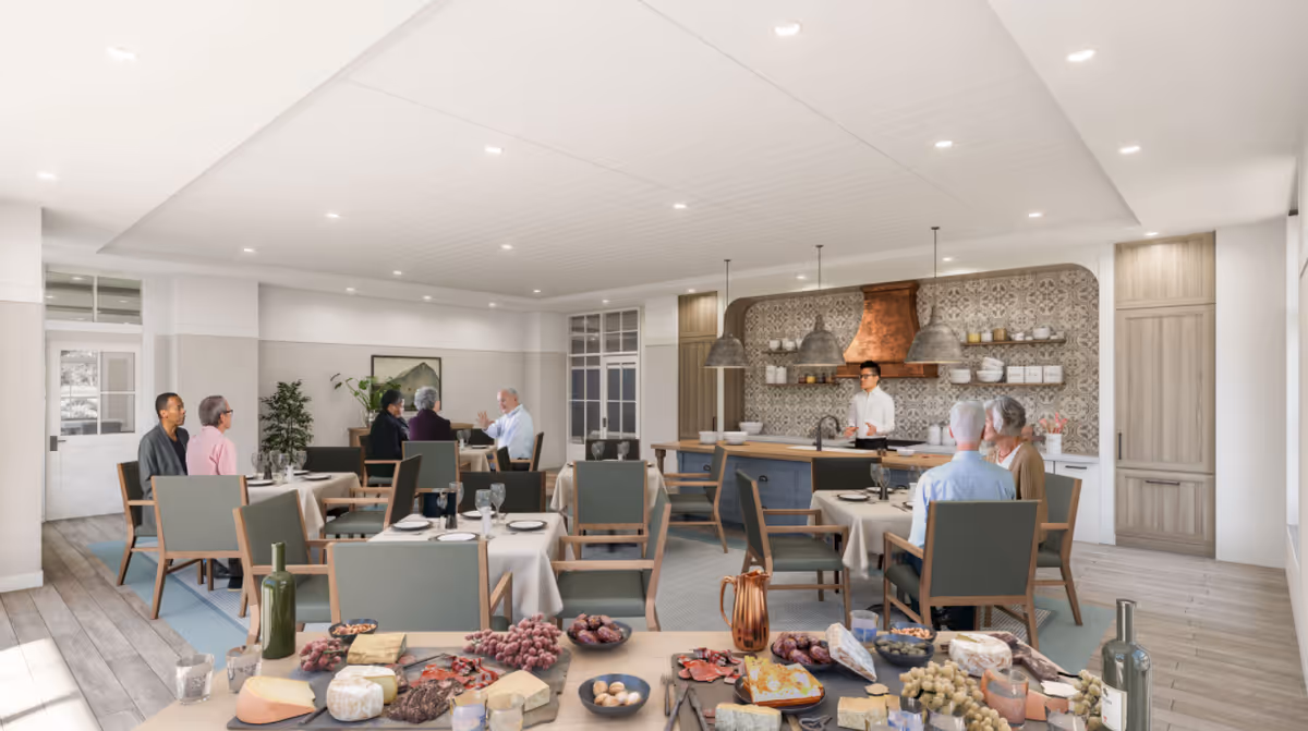 Communal dining room with tables set for meals, residents seated, a food buffet in the foreground, and an open kitchen in the background.